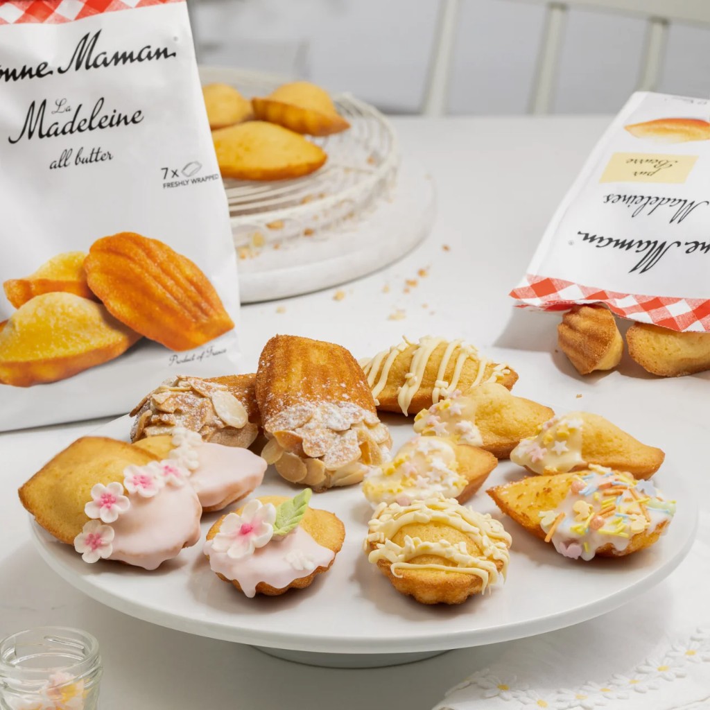 Decorated madeleines on a white cake stand with bakery bags in the background