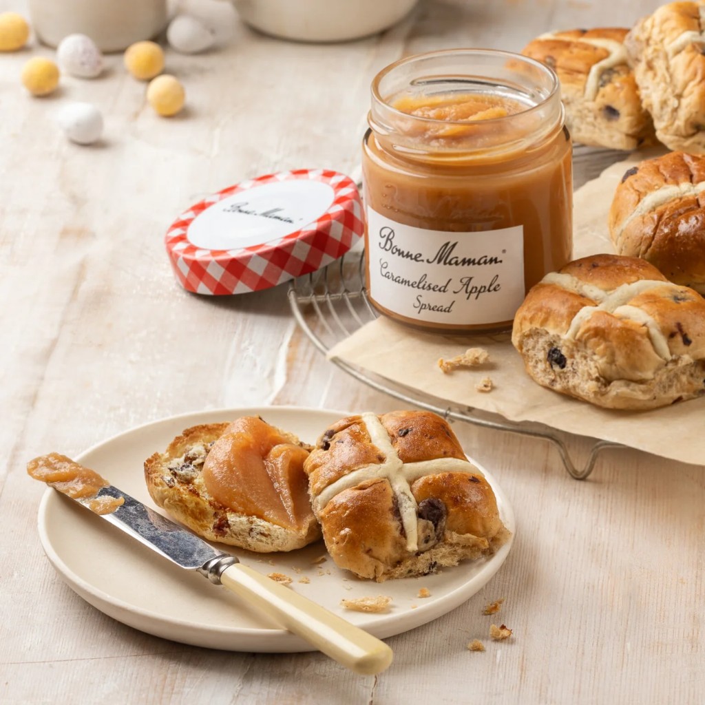 Open jar of caramelised apple spread beside glazed hot cross buns on wood